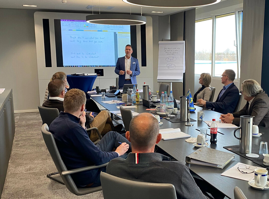 Mehrere Leute sitzen an einem langen Tisch und einer hält eine Präsentation.