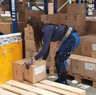 Frau mit einem angelegten Exoskelett hebt Kisten von einer Holzpalette in einem Lager