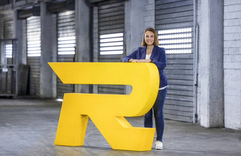 Frau lehnt an großem gelbem Buchstaben R in einer Industriehalle.