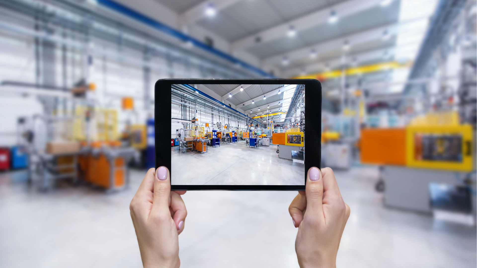 Person hält Tablet mit Blick auf eine moderne Fabrikhalle mit Maschinen und Arbeitsbereichen.