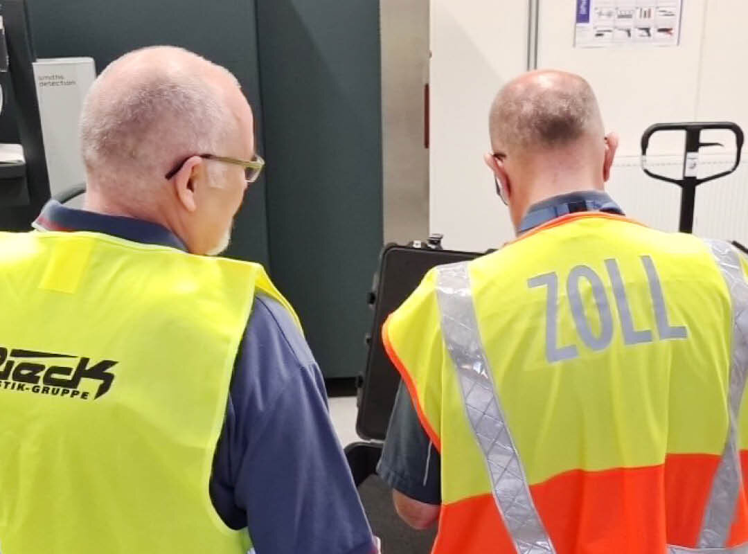 Zwei Männer in Warnwesten, einer mit der Aufschrift "ZOLL", von hinten in einem Innenraum.