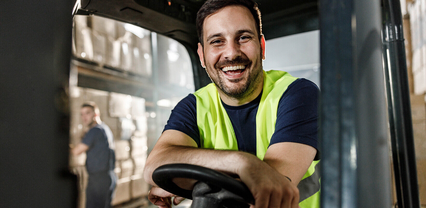 Lächelnder Mann mit Warnweste sitzt auf Gabelstapler in Lagerhalle.