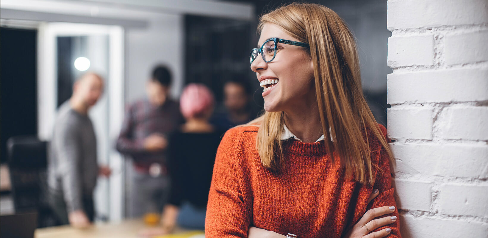 Frau mit Brille und orangem Pullover lehnt lächelnd an einer weißen Wand, im Hintergrund unscharfe Personen in einem Büro.