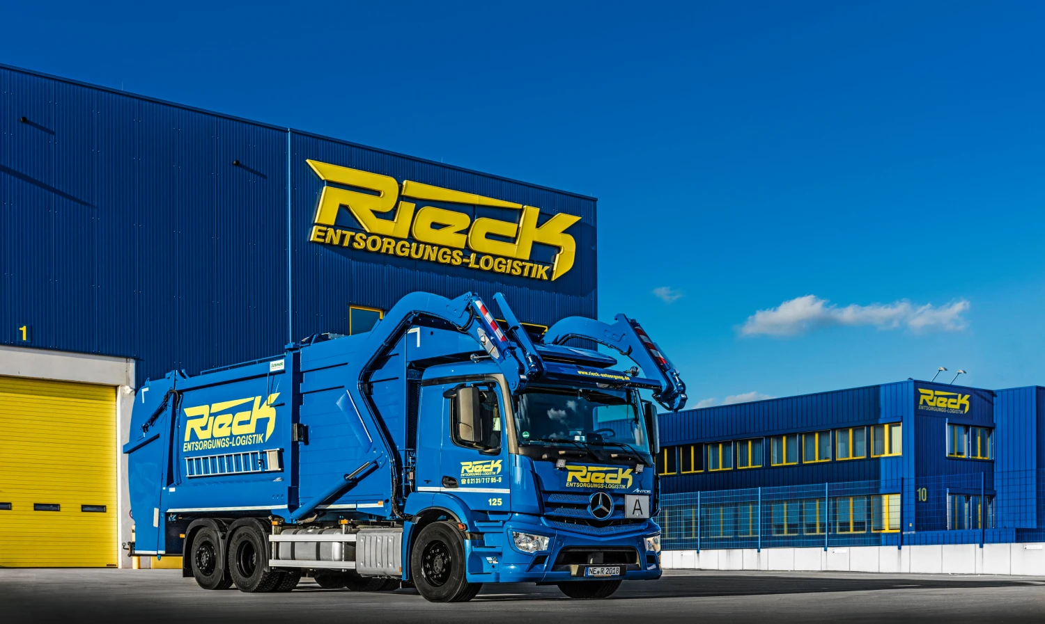 Blauer Müllwagen vor blauem Gebäude mit gelbem Schriftzug Rieck Entsorgungs-Logistik bei klarem Himmel.